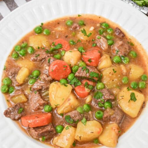 A white bowl filled with Dutch Oven Beef Stew featuring chunks of beef, potatoes, carrots, and green peas, garnished with chopped herbs. Slices of bread and parsley are in the background.