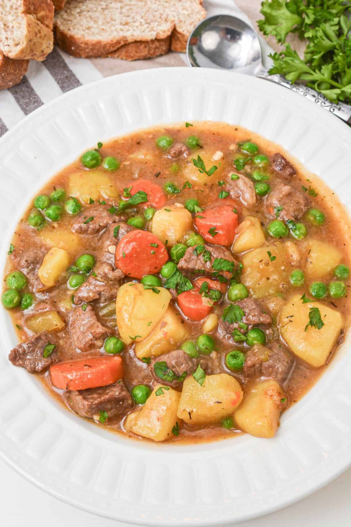 A white bowl filled with Dutch Oven Beef Stew featuring chunks of beef, potatoes, carrots, and green peas, garnished with chopped herbs. Slices of bread and parsley are in the background.