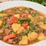 A bowl of Dutch Oven Beef Stew with chunks of beef, carrots, potatoes, and peas in a rich brown broth, garnished with fresh herbs.