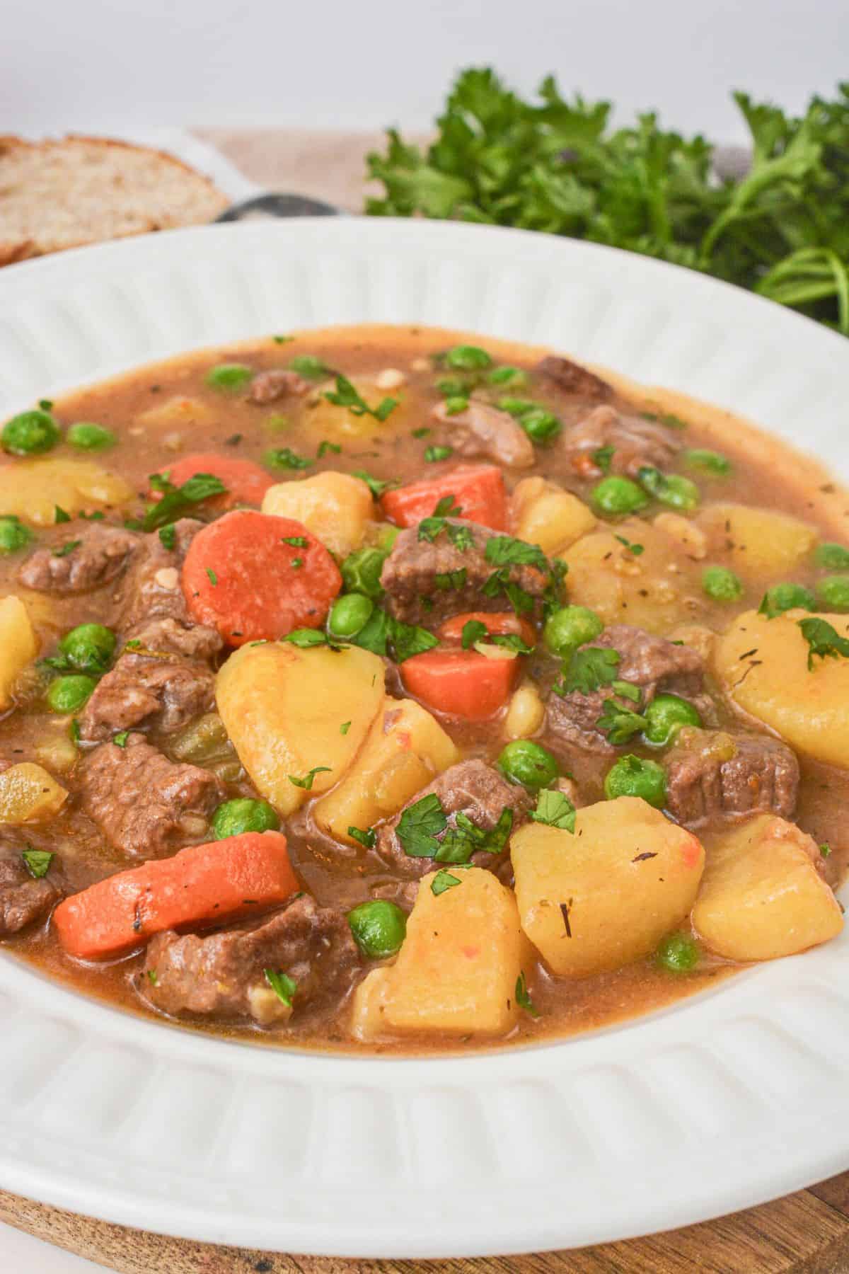 A bowl of Dutch Oven Beef Stew with chunks of beef, carrots, potatoes, and peas in a rich brown broth, garnished with fresh herbs.