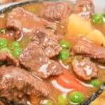 Close-up of a ladle holding chunks of meat, potatoes, carrots, and green peas in a thick, hearty broth.