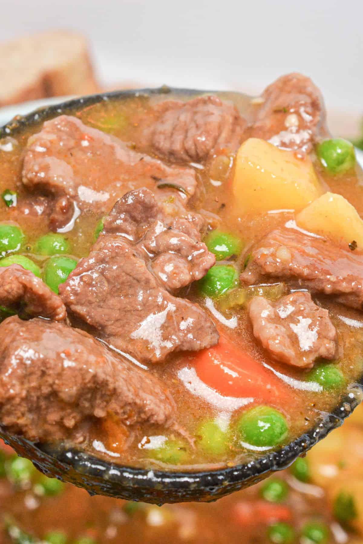 Close-up of a ladle holding chunks of meat, potatoes, carrots, and green peas in a thick, hearty broth.