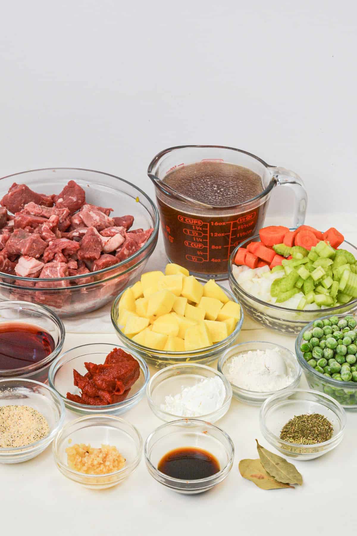 A variety of ingredients including cubed meat, diced potatoes, carrots, celery, onions, peas, broth, seasonings, sauces, tomato paste, garlic, and flour—all arranged in bowls.