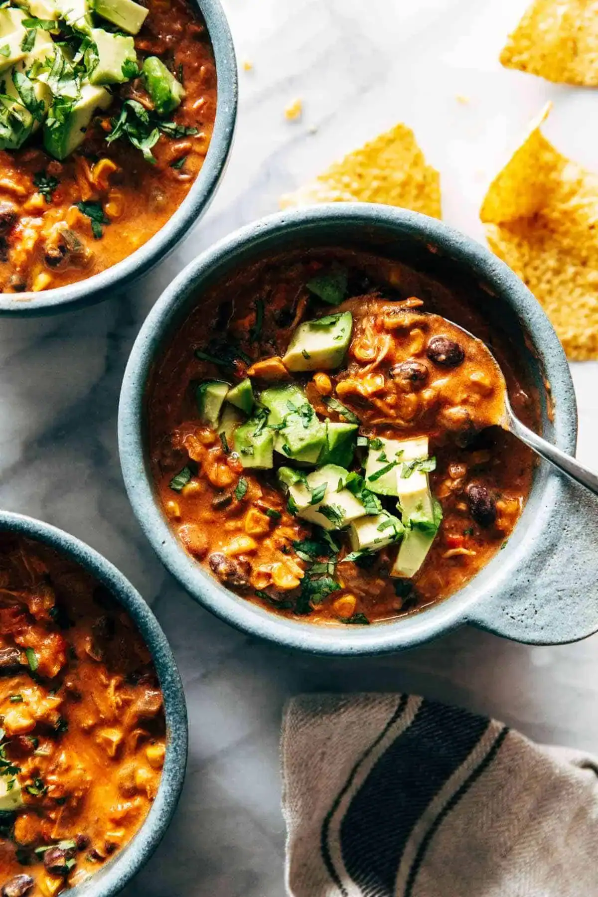 Three bowls of hearty chili—one of those must try chili recipes everyone loves—are topped with diced avocado and herbs, served with tortilla chips on a marble surface. A spoon is in one bowl, and a striped cloth napkin is nearby.