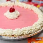 A vibrant Kool Aid Pie with a pink filling, whipped cream border, and a cherry on top is displayed next to colorful Kool-Aid packets and an extra cherry.