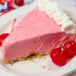 A slice of bright pink Kool Aid Pie with a graham cracker crust, topped with whipped cream and served with maraschino cherries on a white plate.