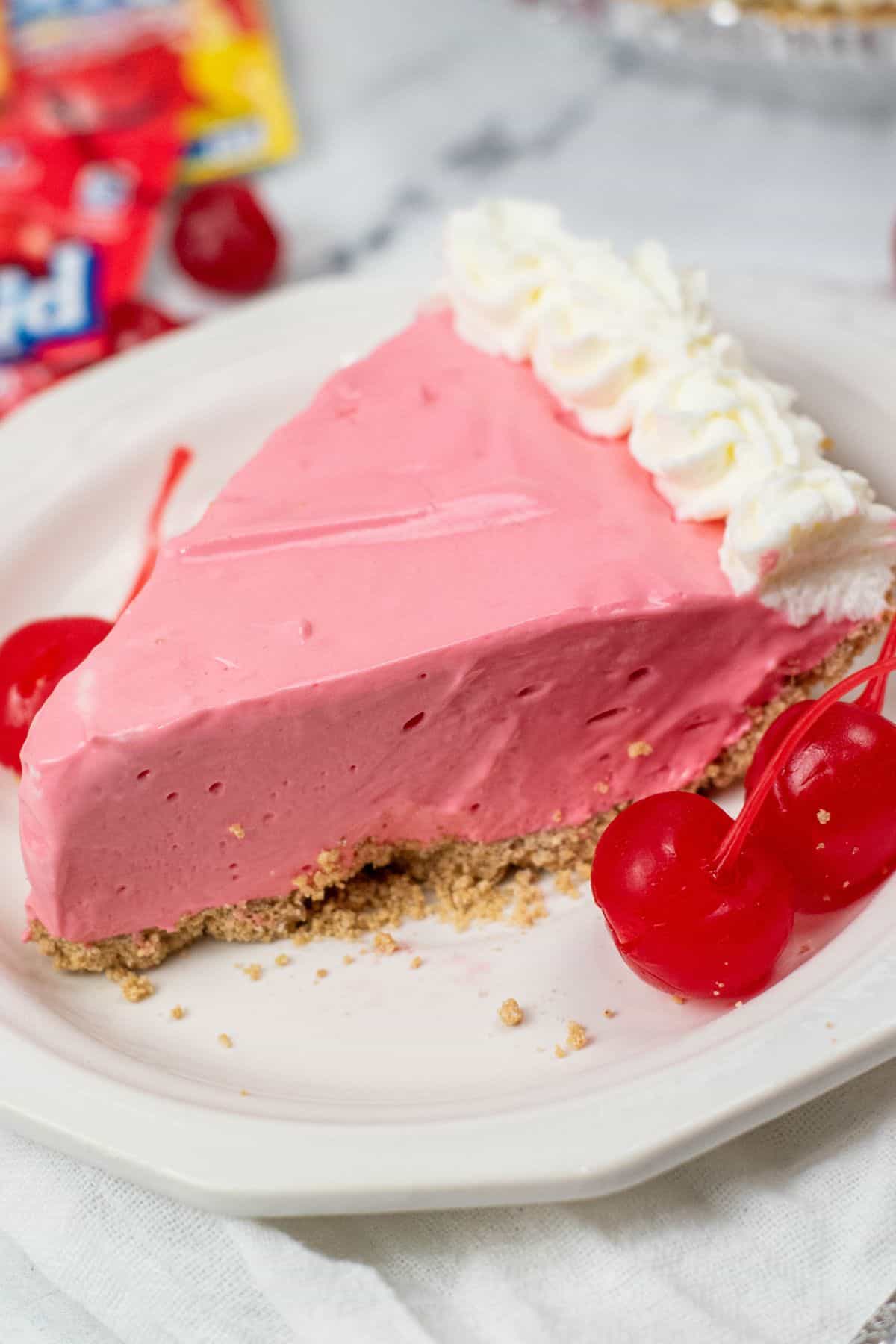 A slice of bright pink Kool Aid Pie with a graham cracker crust, topped with whipped cream and served with maraschino cherries on a white plate.