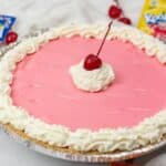 A pink dessert in a foil crust, topped with whipped cream and a cherry; with fresh cherries are visible in the background.