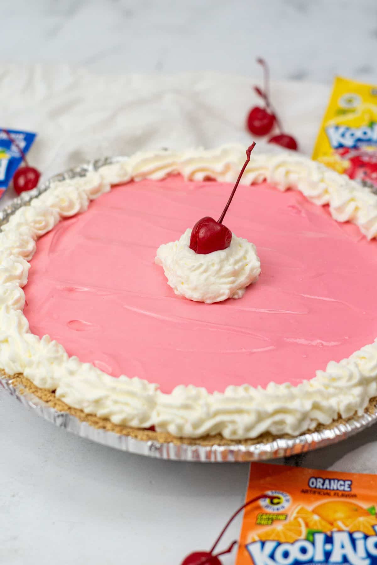 A pink dessert in a foil crust, topped with whipped cream and a cherry; with fresh cherries are visible in the background.