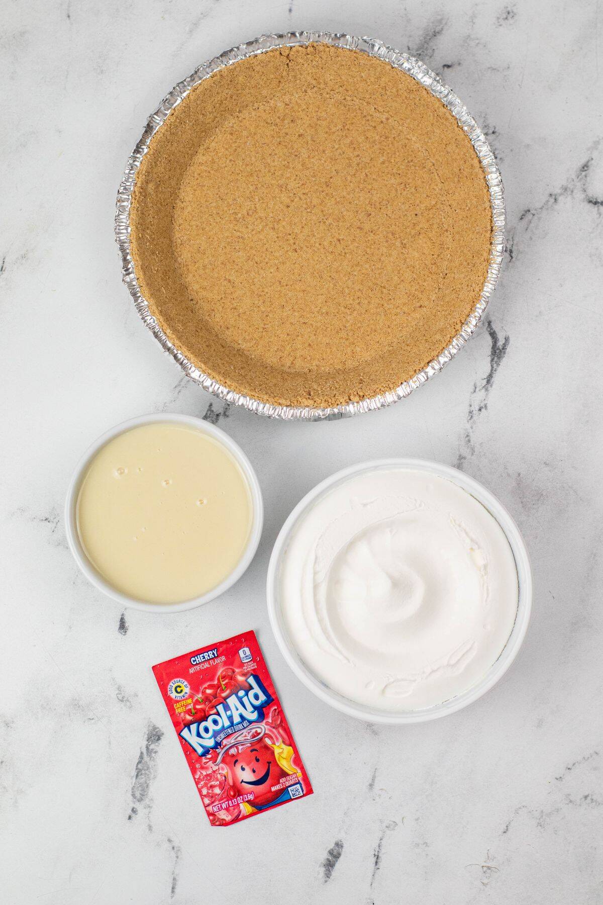 A graham cracker pie crust, a bowl of sweetened condensed milk, a bowl of whipped topping, and a packet of cherry Kool-Aid are ready to come together for a classic Kool Aid Pie on a white marble surface.