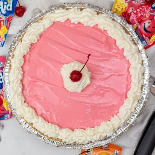 A Kool Aid Pie with a pink cream filling, topped with whipped topping and a cherry in the center, surrounded by cherry and lemonade Kool-Aid packets and loose cherries on a marble surface.