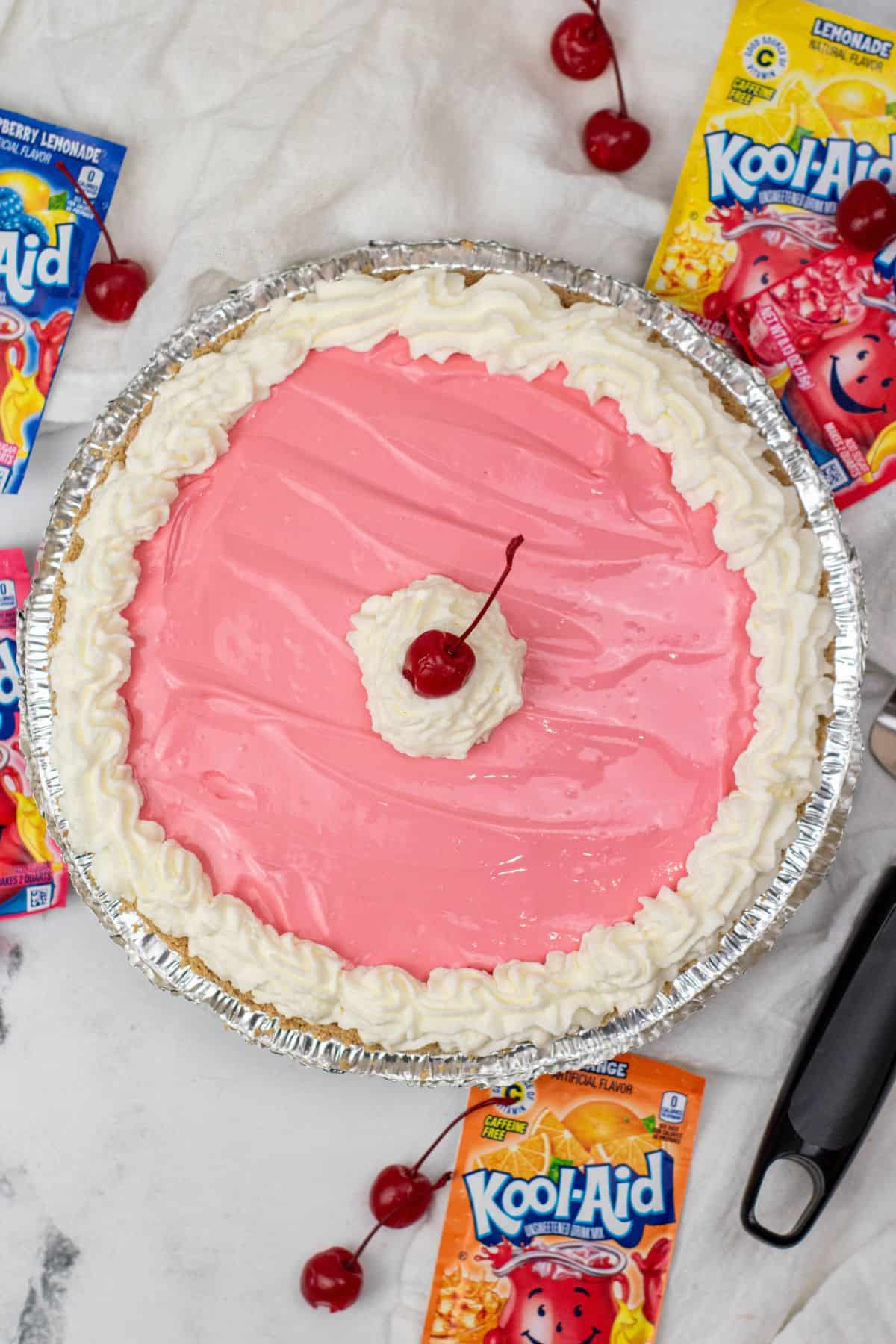 A Kool Aid Pie with a pink cream filling, topped with whipped topping and a cherry in the center, surrounded by cherry and lemonade Kool-Aid packets and loose cherries on a marble surface.
