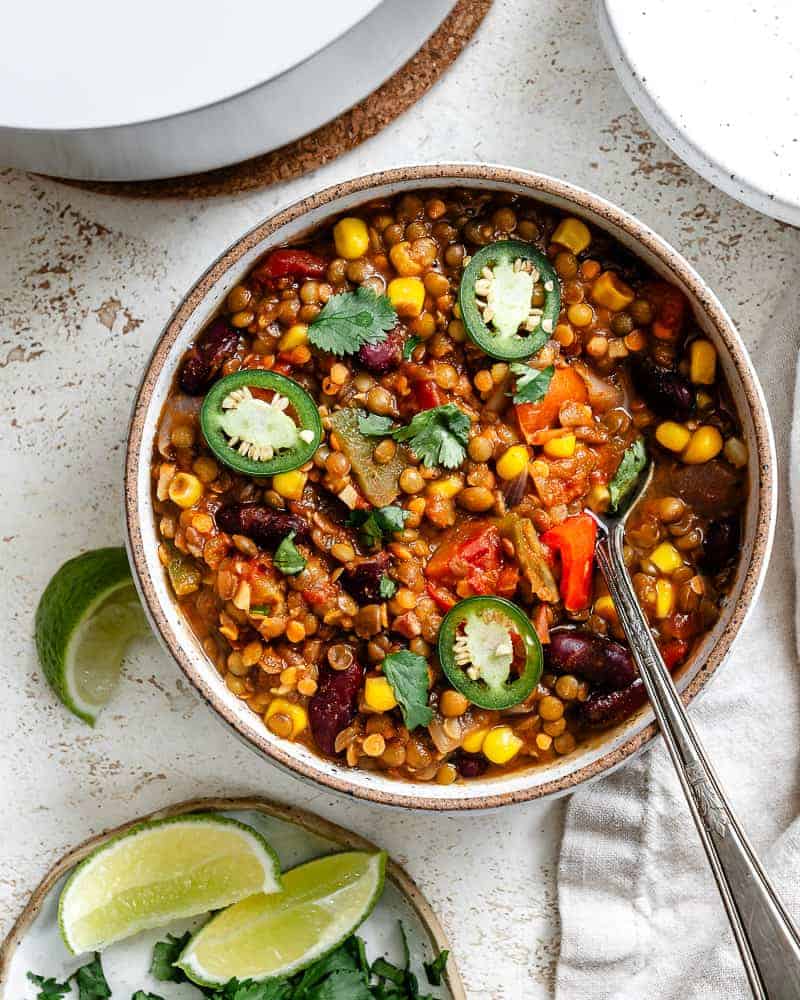 Bowl of lentil chili topped with jalapeño slices and cilantro, with a spoon in the bowl. Lime wedges and chopped cilantro on the side—this is truly a must try for anyone who enjoys hearty chili recipes that everyone loves.