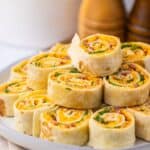 A plate of Pepper Jelly Pinwheels filled with cheese, bacon, and green onions is stacked on a white plate, with salt and pepper shakers in the background.