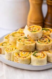 A plate of Pepper Jelly Pinwheels filled with cheese, bacon, and green onions is stacked on a white plate, with salt and pepper shakers in the background.