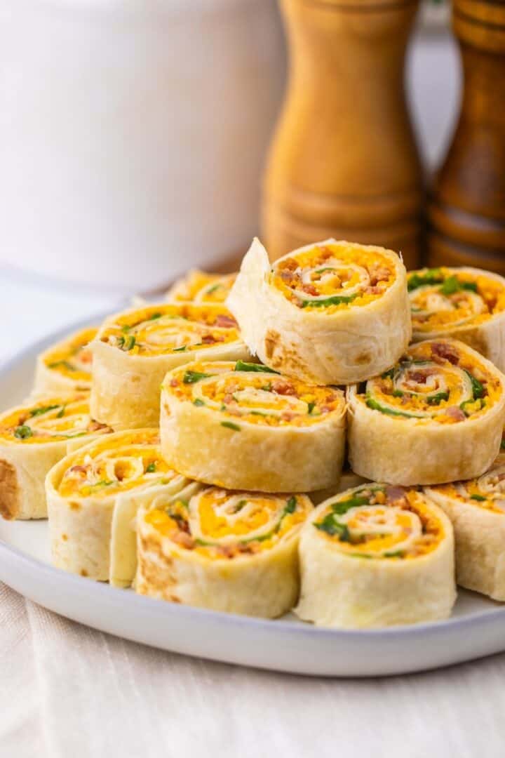 A plate of Pepper Jelly Pinwheels filled with cheese, bacon, and green onions is stacked on a white plate, with salt and pepper shakers in the background.
