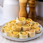A plate of Pepper Jelly Pinwheels filled with deli meat, cheese, and greens sits on a table with salt and pepper shakers and a potted plant in the background.