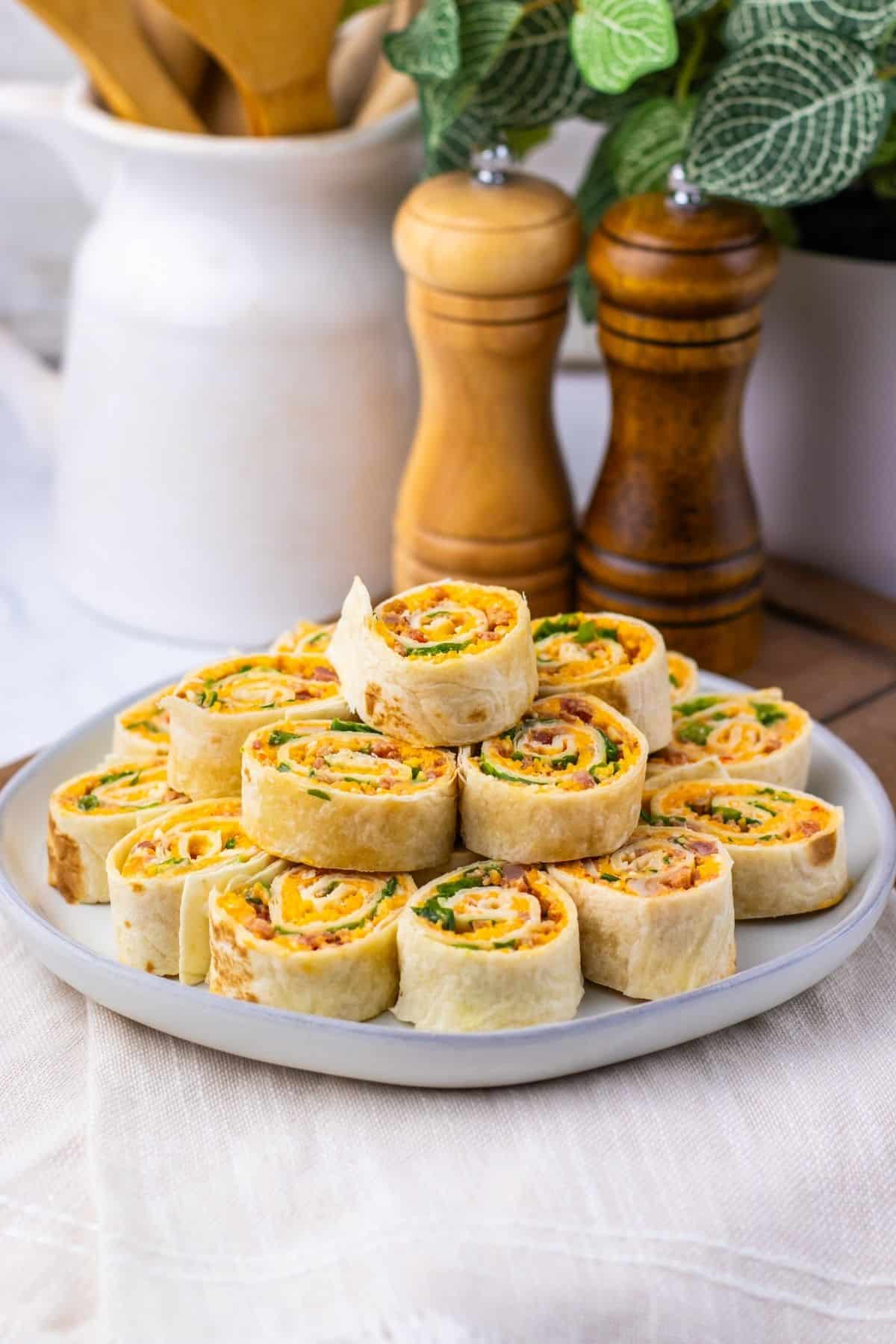A plate of Pepper Jelly Pinwheels filled with deli meat, cheese, and greens sits on a table with salt and pepper shakers and a potted plant in the background.