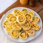 A plate of Pepper Jelly Pinwheels filled with cheese, greens, and other ingredients is arranged neatly on a white plate, with salt and pepper shakers in the background.