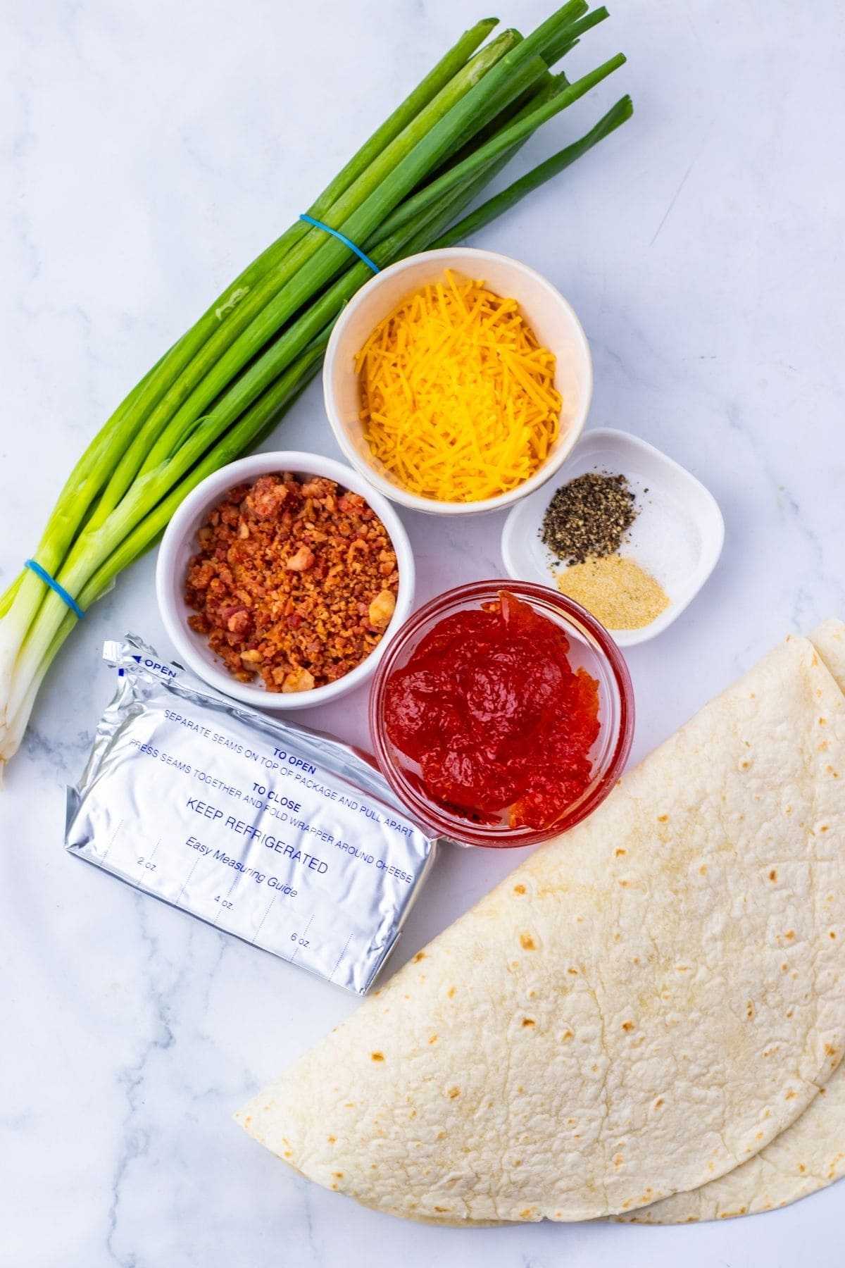 Ingredients on a marble surface  green onions, shredded cheddar cheese, bacon bits, shredded cheese, spices, tomato sauce, cream cheese, and flour tortillas.