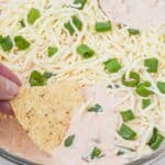 A hand dips a tortilla chip into a glass dish topped with shredded cheese and chopped green onions.