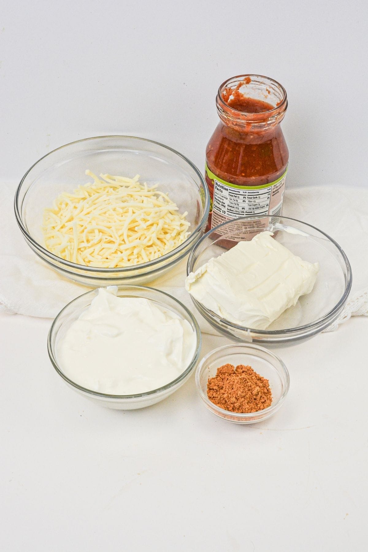 Five ingredients are displayed for Salsa and Cream Cheese Dip: shredded cheese, a jar of tomato sauce, a block of cream cheese, a bowl of sour cream, and a small bowl of seasoning mix.