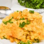 A serving of Slow Cooker Corn Casserole topped with chopped parsley on a white plate, with a spoon and fresh parsley in the background.