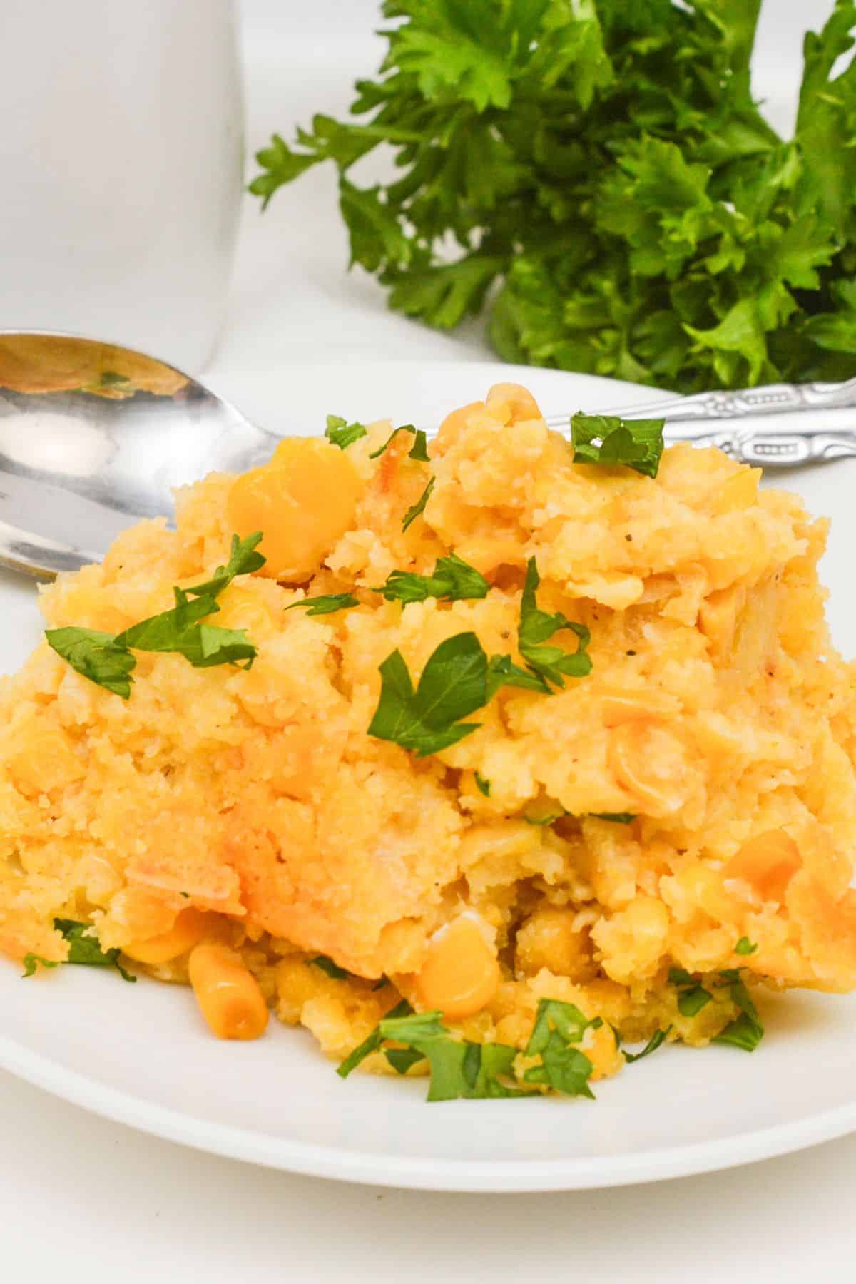 A serving of Slow Cooker Corn Casserole topped with chopped parsley on a white plate, with a spoon and fresh parsley in the background.