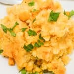 A serving of Slow Cooker Corn Casserole garnished with chopped parsley on a white plate, with a metal spoon in the background.