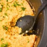 A black spoon rests in a slow cooker with Slow Cooker Corn Casserole, its golden top sprinkled with chopped herbs and a hearty portion already served.