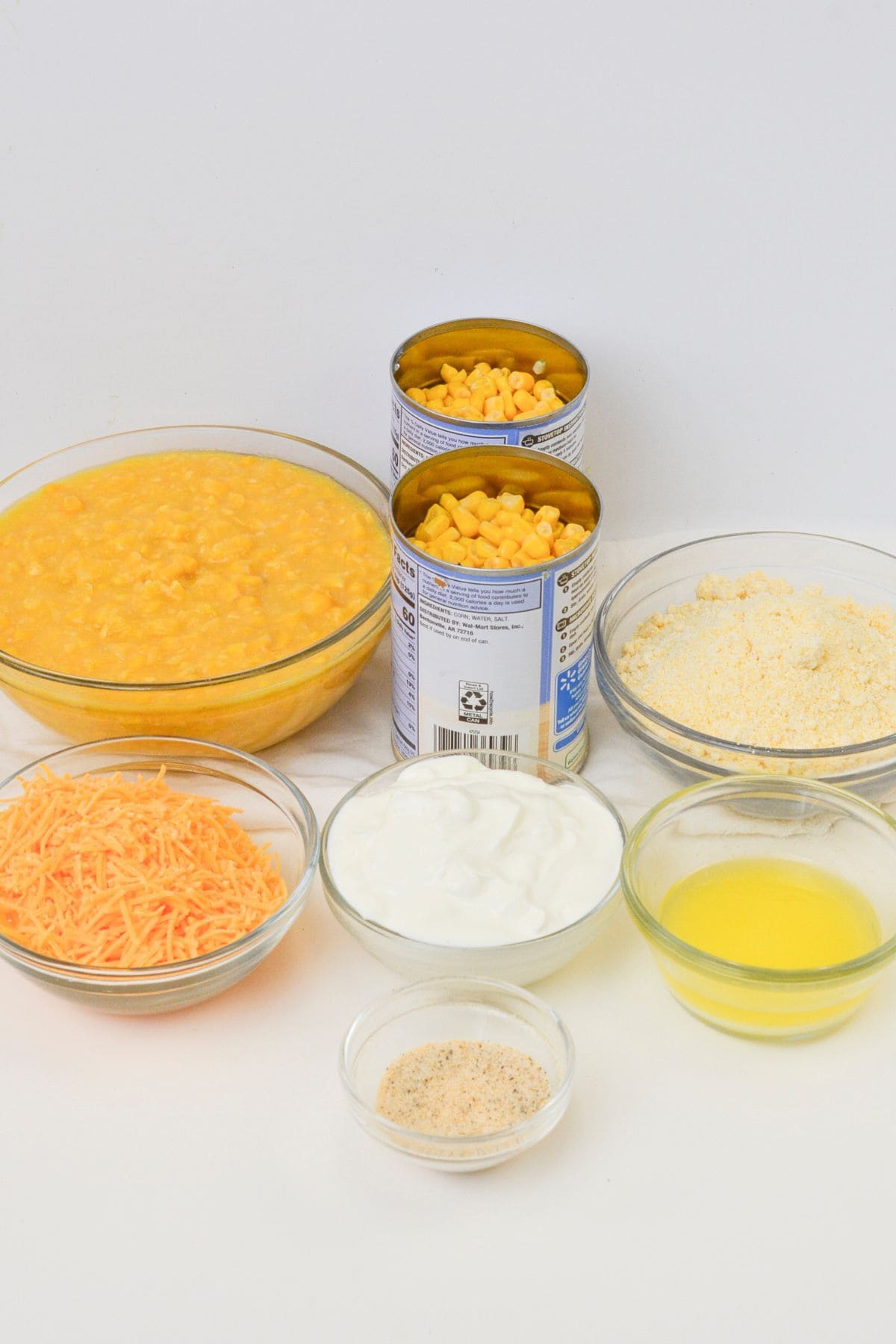 Various ingredients for Slow Cooker Corn Casserole, including creamed corn, canned corn, shredded cheese, cornmeal, sour cream, melted butter, and breadcrumbs, are displayed in bowls on a white background.