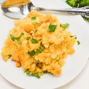 A serving of Slow Cooker Corn Casserole topped with chopped parsley on a white plate, with a silver spoon and fresh herbs beside it.