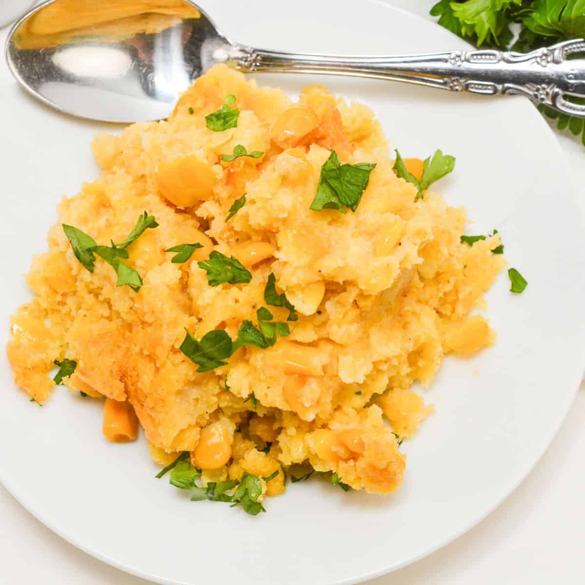A serving of Slow Cooker Corn Casserole topped with chopped parsley on a white plate, with a silver spoon and fresh herbs beside it.