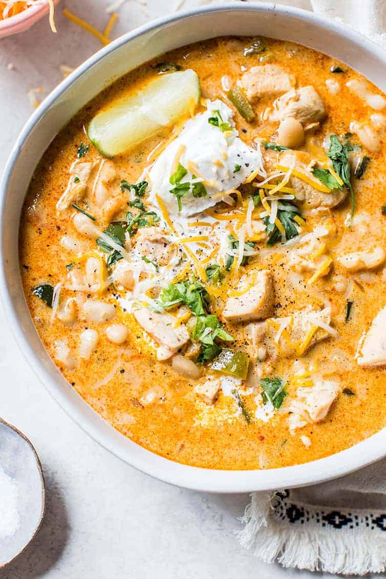 Bowl of creamy chicken chili—one of those must try chili recipes—topped with shredded cheese, sour cream, chopped herbs, and a lime wedge that everyone loves.