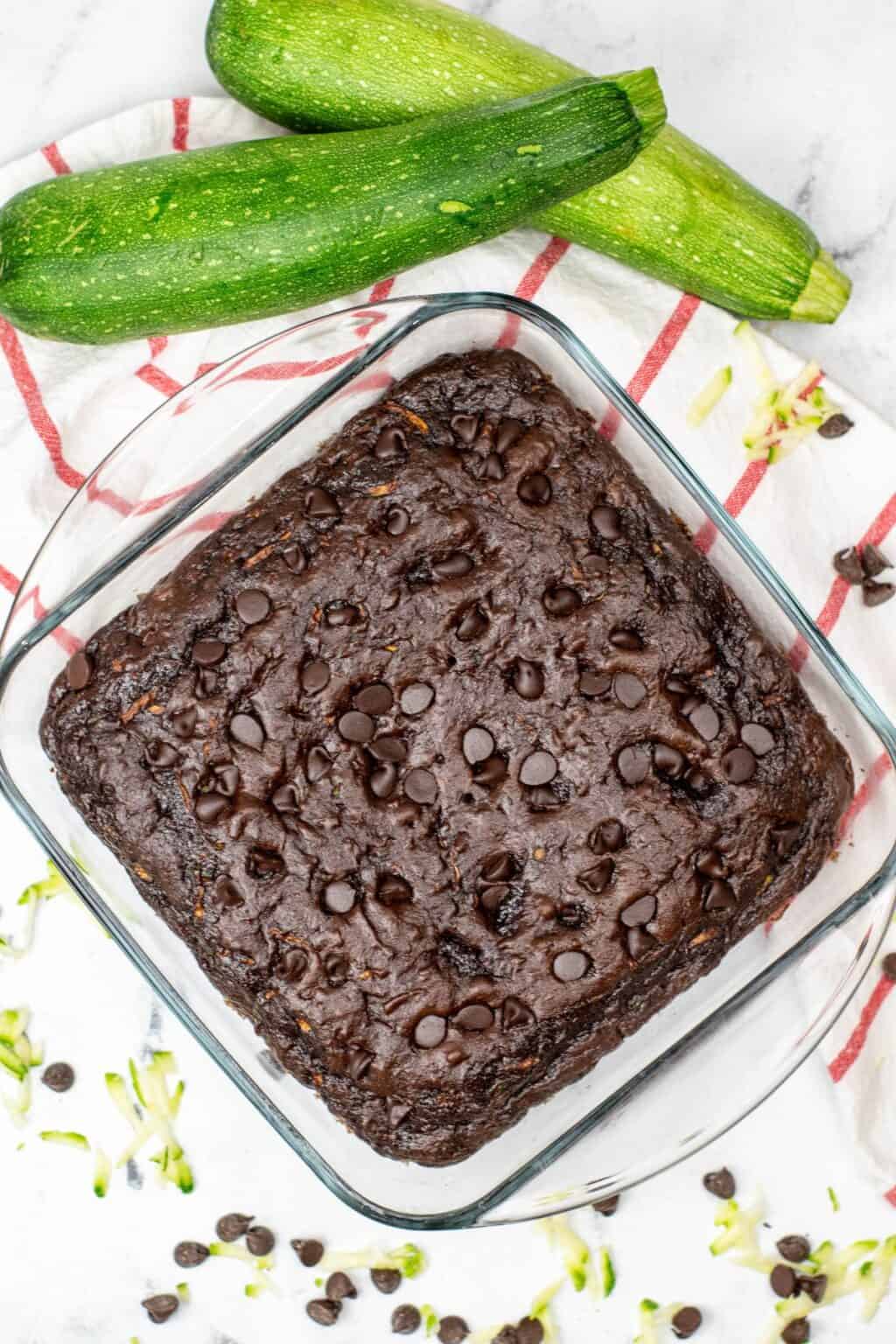 A glass baking dish with rich zucchini chocolate brownies, reminiscent of Zucchini Chocolate Cake, topped with chocolate chips, sits on a red-striped towel beside two whole zucchinis.