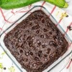 A glass baking dish with rich zucchini chocolate brownies, reminiscent of Zucchini Chocolate Cake, topped with chocolate chips, sits on a red-striped towel beside two whole zucchinis.