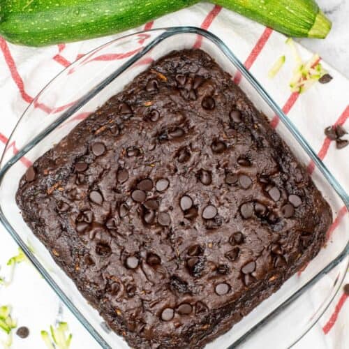A glass baking dish with rich zucchini chocolate brownies, reminiscent of Zucchini Chocolate Cake, topped with chocolate chips, sits on a red-striped towel beside two whole zucchinis.