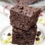 Two pieces of Zucchini Chocolate Cake stacked on a white plate, topped with chocolate chips, with zucchini shreds and more chocolate chips scattered around.