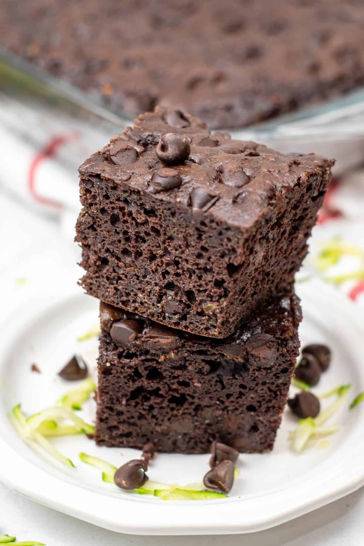 Two pieces of Zucchini Chocolate Cake stacked on a white plate, topped with chocolate chips, with zucchini shreds and more chocolate chips scattered around.