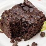 A square piece of Zucchini Chocolate Cake with chocolate chips on top sits on a white plate, with a few chocolate chips and zucchini shreds beside it.