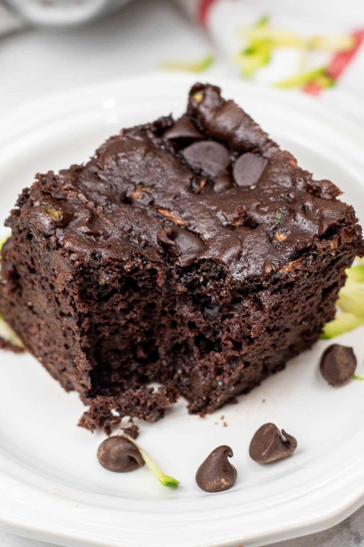 A square piece of Zucchini Chocolate Cake with chocolate chips on top sits on a white plate, with a few chocolate chips and zucchini shreds beside it.
