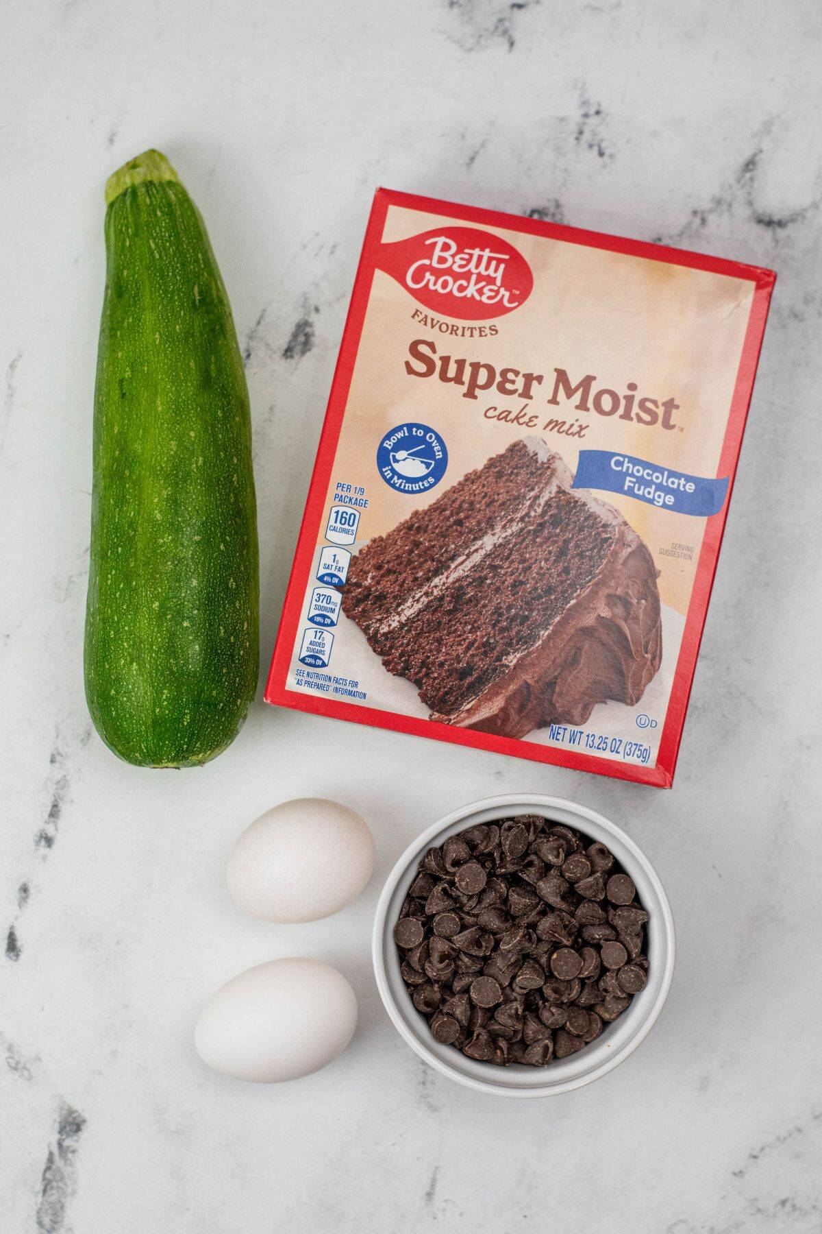 A box  mix, a zucchini, two eggs, and a bowl of chocolate chips on a white surface—perfect for making moist and delicious dessert.