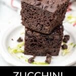 Two pieces of rich Zucchini Chocolate Cake are stacked on a white plate, topped with chocolate chips and surrounded by fresh zucchini shreds.