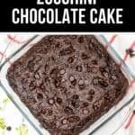 A square glass dish filled with moist Zucchini Chocolate Cake topped with chocolate chips, surrounded by scattered chocolate chips and shredded zucchini.