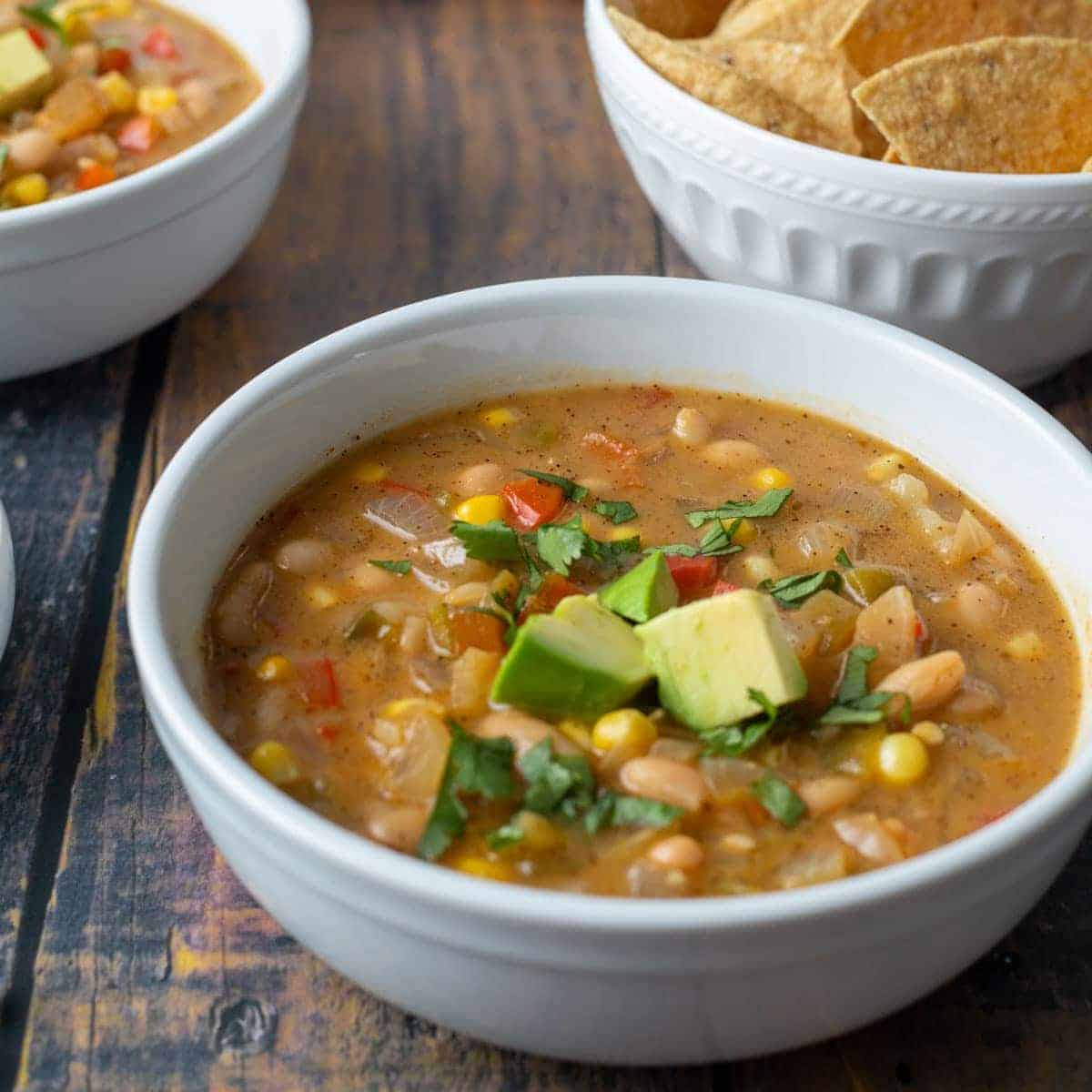 A white bowl of soup containing beans, corn, diced avocado, and herbs on a wooden table, with another bowl of chips in the background—a must try for fans of comforting chili recipes that everyone loves.