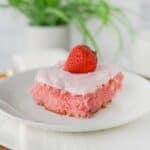 A slice of homemade strawberry cake with white frosting and a whole strawberry on top is served on a white plate with a white napkin; a green plant is in the background.