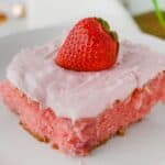 A slice of homemade strawberry cake with pink frosting, topped with a whole fresh strawberry, served on a white plate.