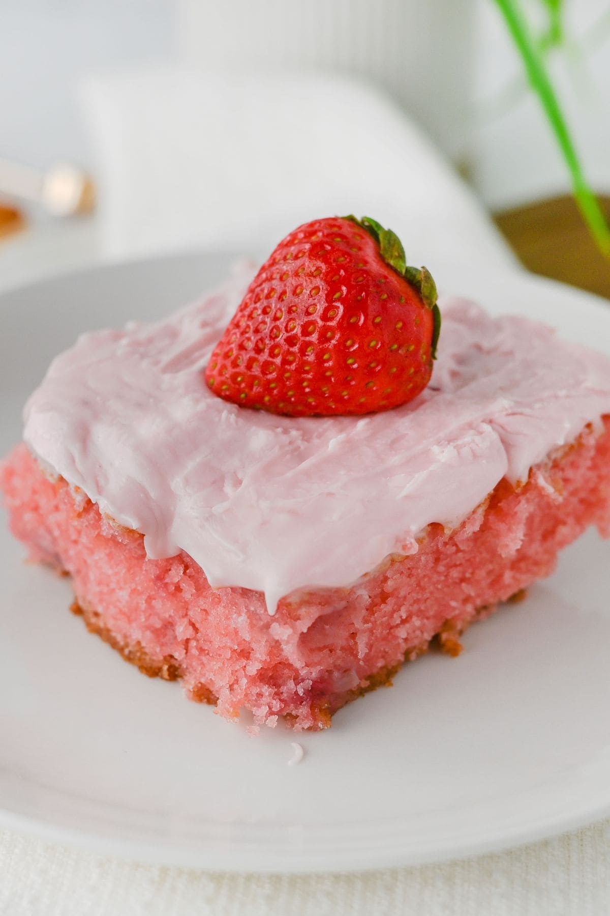 A slice of homemade strawberry cake with pink frosting, topped with a whole fresh strawberry, served on a white plate.