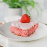 A slice with light pink frosting and a whole strawberry on top, served on a white plate with a white napkin.
