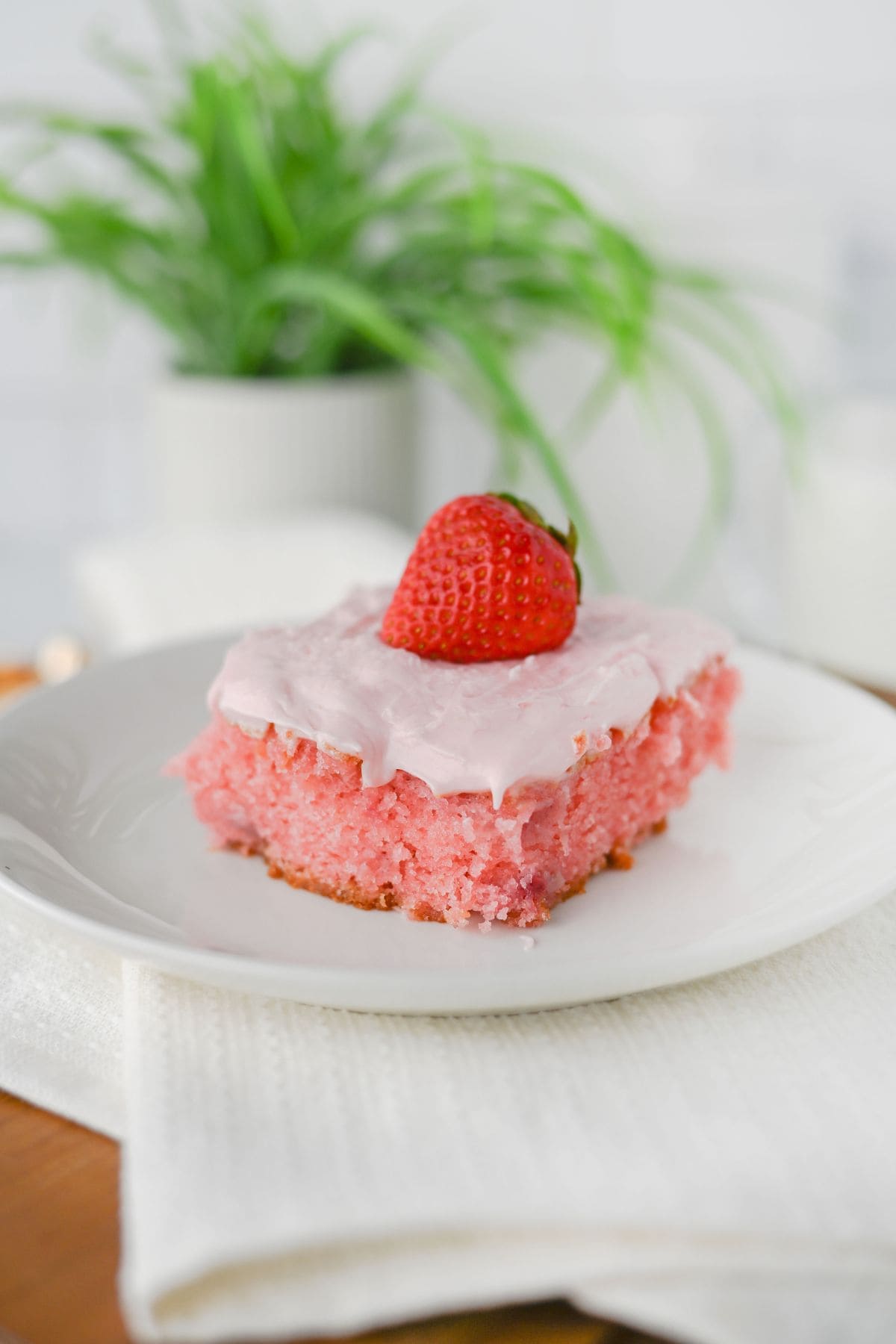 A slice  with light pink frosting and a whole strawberry on top, served on a white plate with a white napkin.